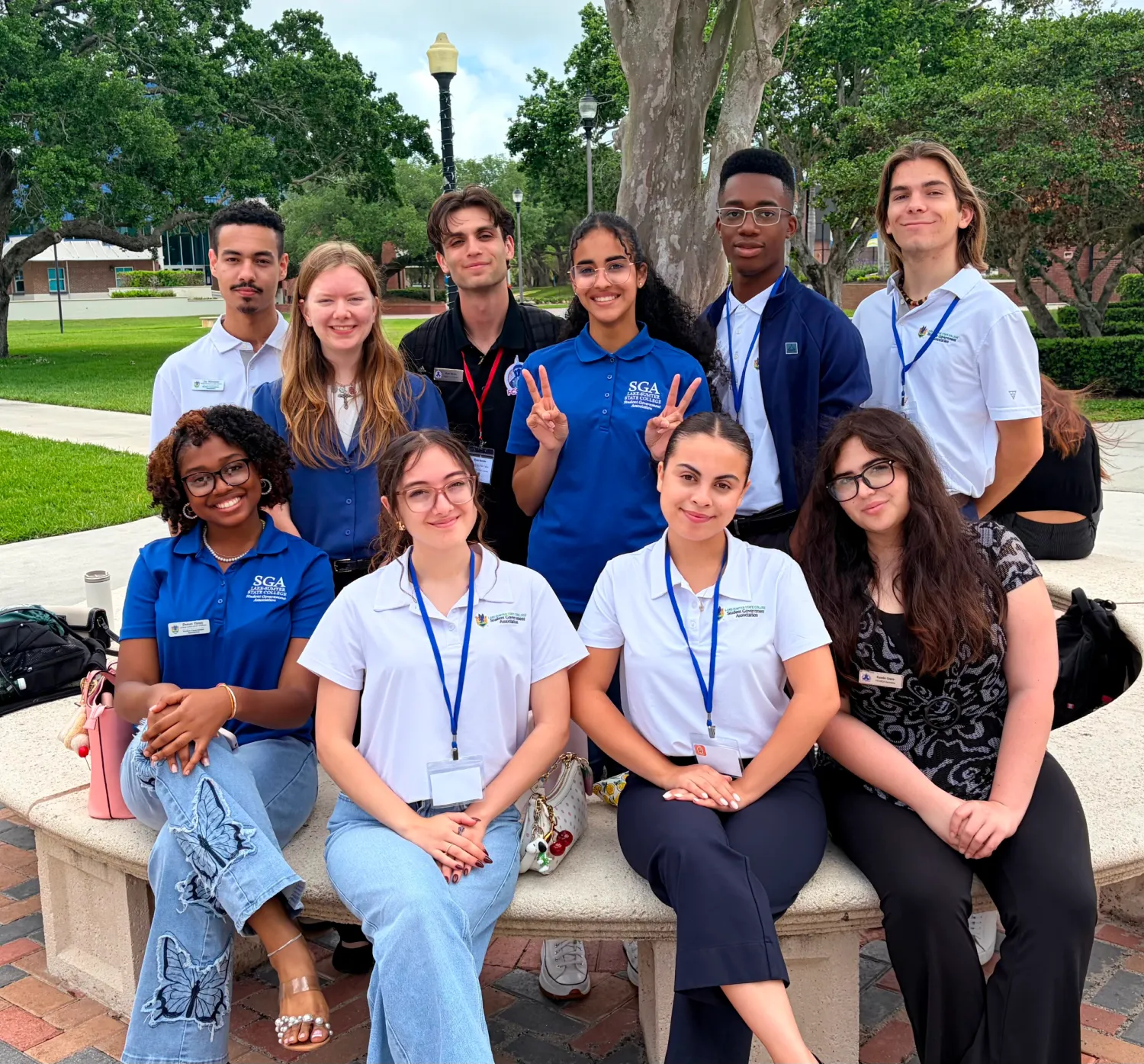 A group of nine SGA students, some in white or blue SGA polo shirts and name tags, sit and stand on a campus bench outdoors, posing for a photo.